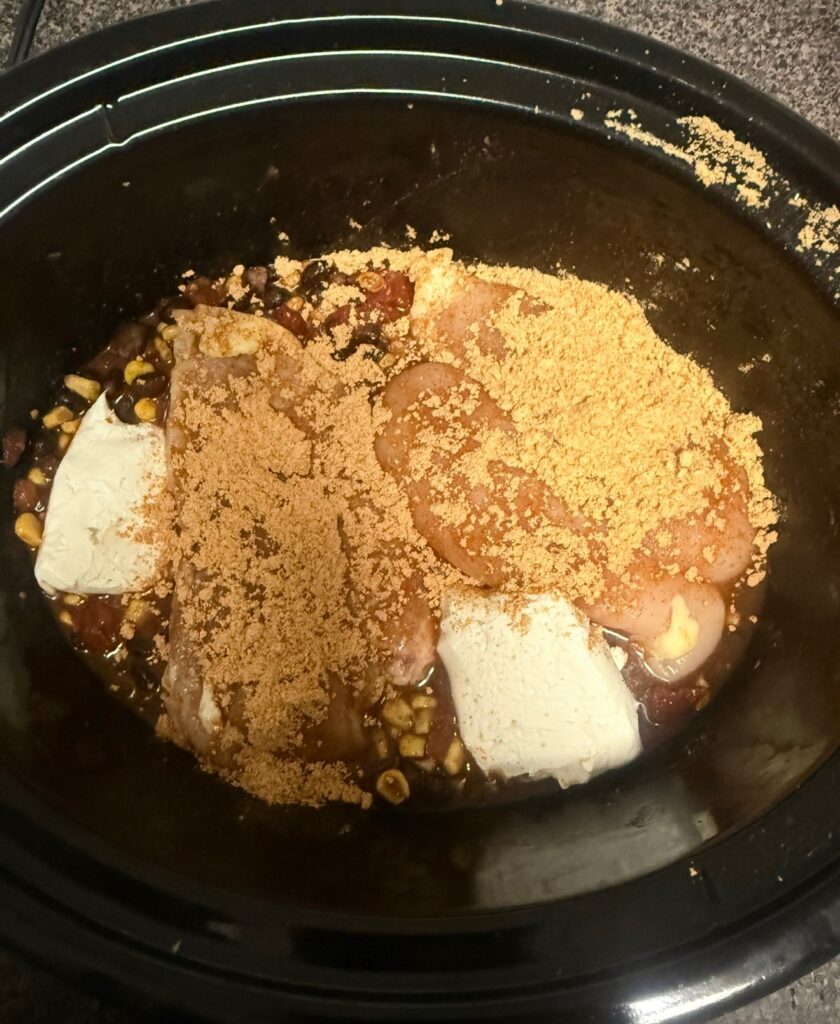 Raw chicken breasts, cream cheese chunks, and seasoning powder in the black crock pot